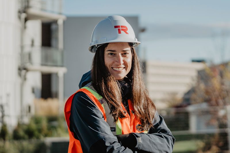 Une femme portant un casque de chantier blanc et un gilet de sécurité orange se tient à l'extérieur sur un chantier, souriante et les bras croisés. Des bâtiments et de la verdure sont visibles à l'arrière-plan.