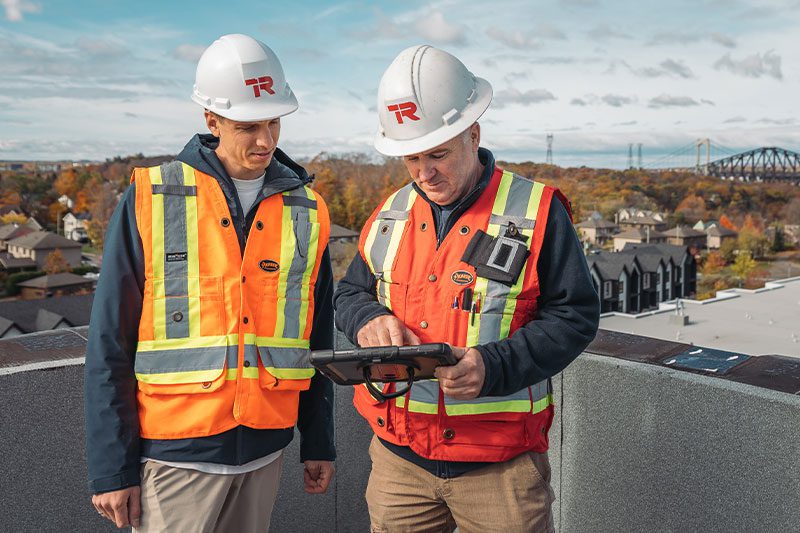 Deux ouvriers du bâtiment portant des gilets de sécurité et des casques se tiennent sur un toit et regardent une tablette. Des maisons, des arbres au feuillage d'automne et un pont sont visibles à l'arrière-plan.
