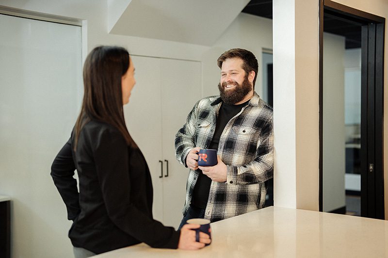 Deux personnes se tiennent dans la cuisine d'un bureau, souriant et tenant des tasses de café tout en discutant. L'un porte une chemise à carreaux et l'autre un blazer noir.