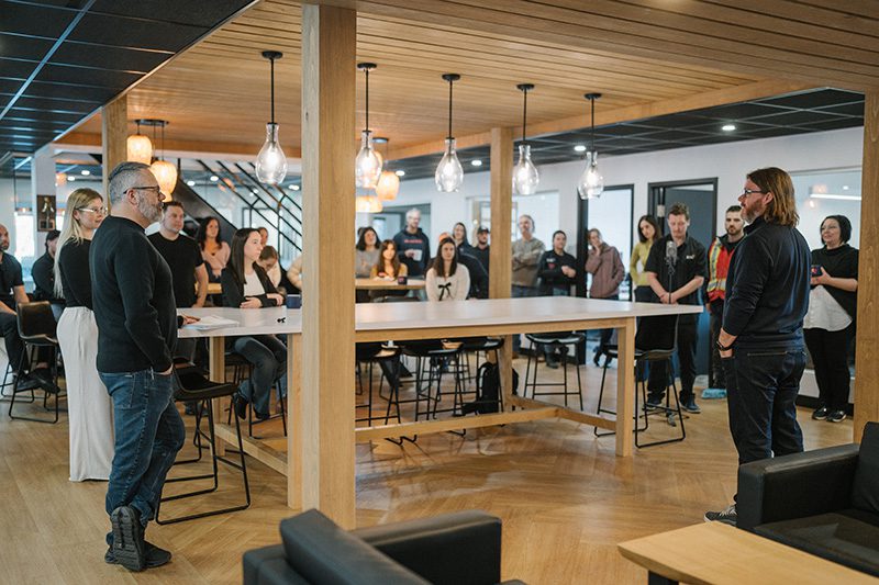 Un groupe de personnes se réunit dans un bureau moderne, écoutant attentivement une personne qui parle à l'avant. La pièce est ornée de bois, de lampes suspendues et d'une grande table autour de laquelle des personnes sont assises ou debout.