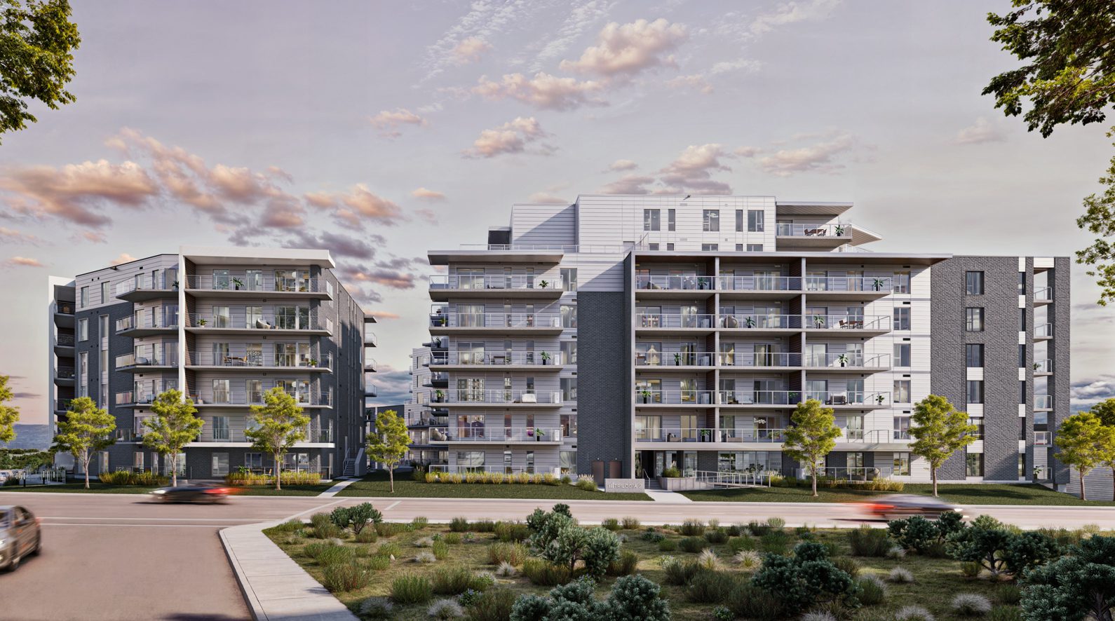 Immeubles modernes de taille moyenne avec de grands balcons, entourés d'arbres et d'aménagements paysagers, situés le long d'une rue où le mouvement des voitures qui passent est flou, sous un ciel partiellement nuageux au coucher du soleil.