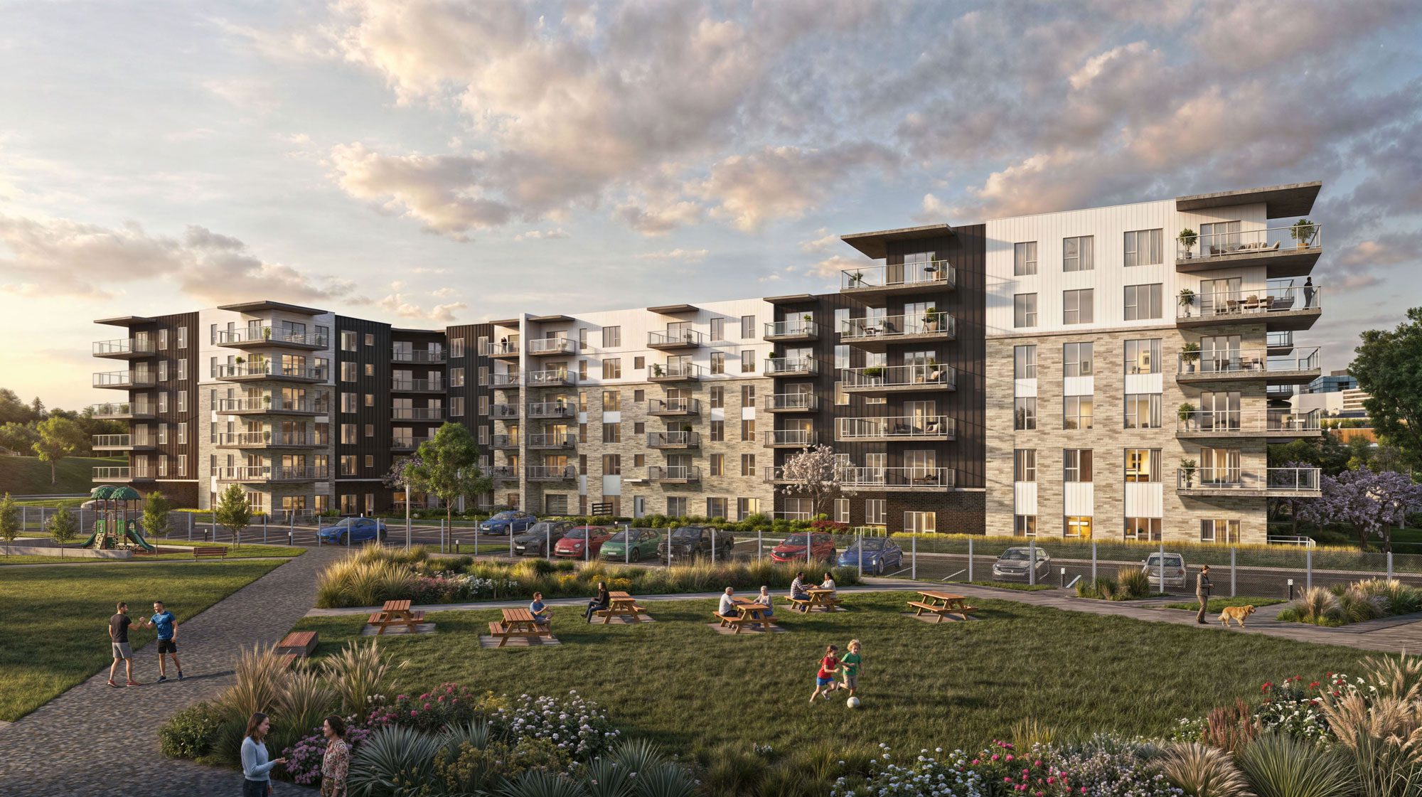 Des immeubles d'habitation modernes avec balcons surplombent un parc verdoyant avec des tables de pique-nique, des gens assis, des enfants qui jouent, une aire de jeux et des voitures garées, le tout sous un ciel partiellement nuageux au coucher du soleil.