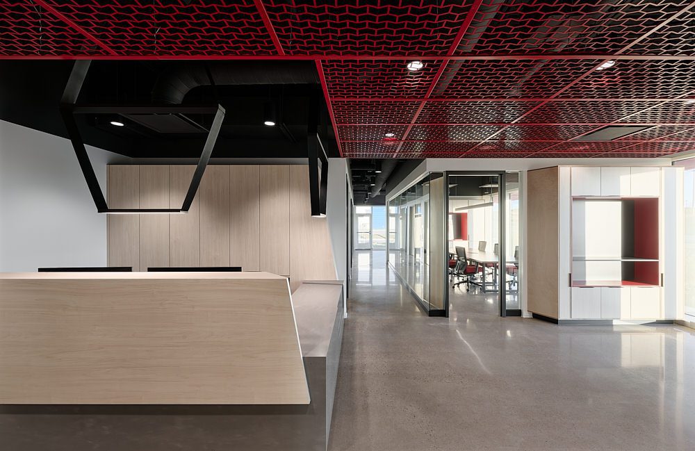 Intérieur de bureau moderne avec un comptoir d'accueil en bois aux formes géométriques, des sols en béton poli, des salles de réunion aux parois vitrées et une ossature de plafond noire rehaussée de touches de rouge, créant ainsi un espace de travail au design épuré et contemporain.