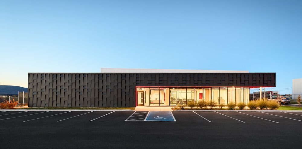 Un bâtiment moderne, de plain-pied, doté d'une façade sombre et texturée, de grandes fenêtres et d'une entrée vitrée. Un parking vide, avec une place réservée aux personnes à mobilité réduite, se trouve devant. Le ciel est dégagé et bleu.