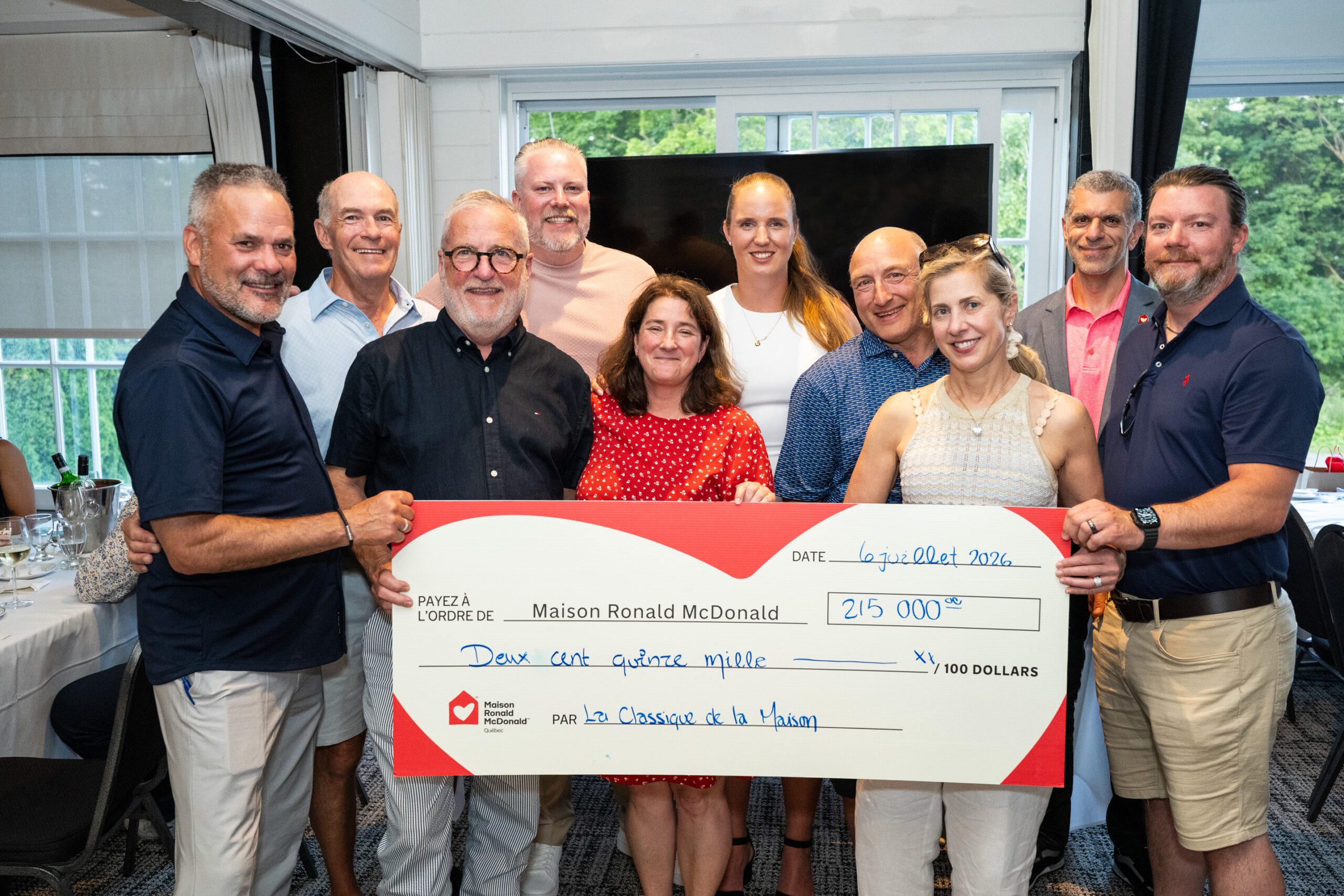 Un groupe de personnes souriant et posant ensemble à l'intérieur, tenant un grand chèque symbolique libellé à l'ordre de la Maison Ronald McDonald d'un montant de 215 000 dollars.