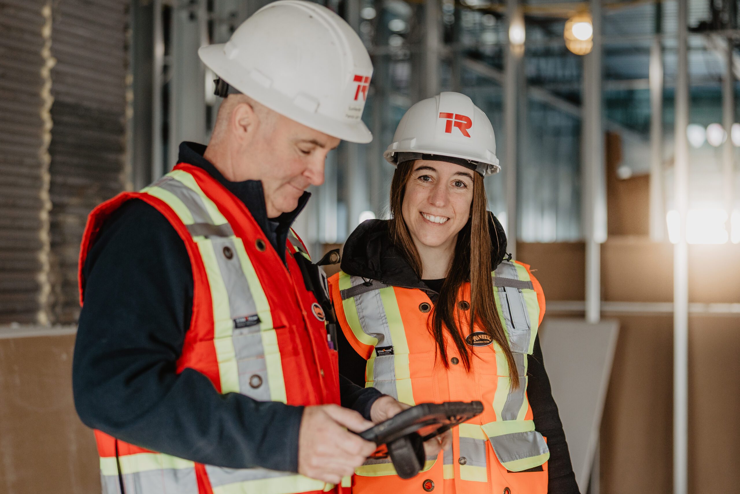 Deux ouvriers du bâtiment, coiffés de casques blancs et vêtus de gilets réfléchissants, se tiennent à l'intérieur d'un chantier. L'un d'eux consulte une tablette, tandis que l'autre sourit à l'appareil photo. On aperçoit en arrière-plan une ossature métallique et des murs en cours de finition.